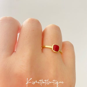 May include: A gold ring featuring a square, red gemstone. The ring is worn on a finger, set against a plain white backdrop. The ring's design is simple, with a slender band and a bezel setting for the red gemstone. The image includes the text "Kismetkatboutique".
