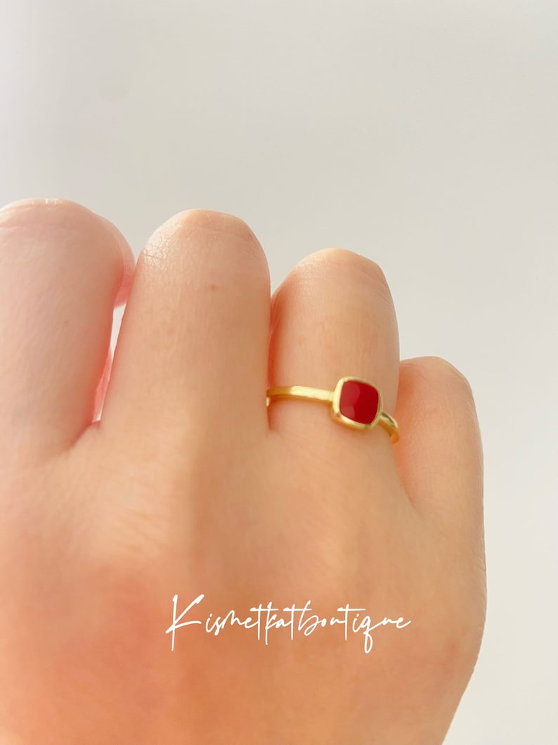 May include: A gold-colored ring with a square, red gemstone. The ring is on a finger, and the background is a plain white. The ring is a simple design, with a thin band and a bezel setting for the gemstone. The text "Kismetkatboutique" is in the image.