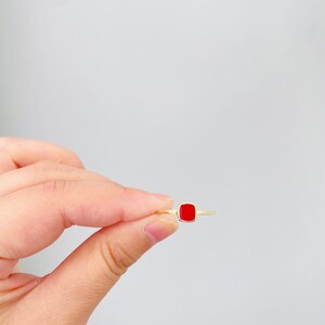 May include: A gold ring featuring a square, bright red gemstone. The ring is held between fingers against a white background, showcasing the minimalist design and vibrant color of the jewelry.