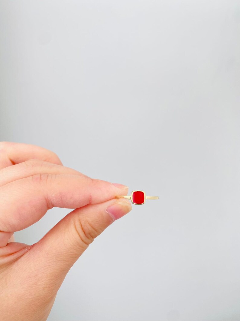 May include: A gold ring featuring a square, bright red gemstone. The ring is held between fingers against a white background, showcasing the minimalist design and vibrant color of the jewelry.