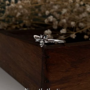 Op de afbeelding: Een zilverkleurige ring met een gedetailleerd bijenontwerp. De bij heeft getextureerde vleugels en lichaamssegmenten. De ring ligt op een donkere houten ondergrond met de tekst "Kismetkatboutique".