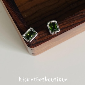 May include: A pair of stud earrings featuring rectangular, deep green faceted gemstones set in polished silver-toned metal frames. The earrings are positioned near the corner of a dark wood box. The text Kismetkatboutique is visible below.