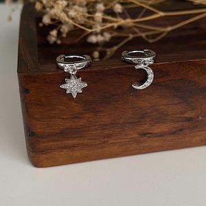 May include: Silver hoop earrings with celestial charms. One earring features a starburst design, the other a crescent moon, both adorned with small, sparkling stones. The earrings are displayed on a wooden surface.