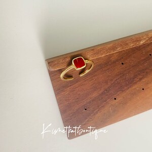 May include: A gold-toned ring with an open design, featuring a square, vibrant red stone. The ring is displayed on a wooden surface. The text "Kismetkatboutique" is visible in the image.