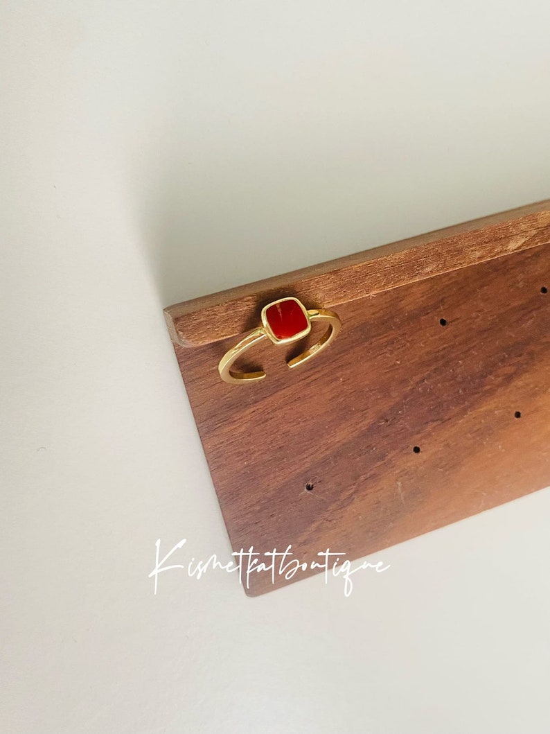 May include: A gold-toned ring with an open design, featuring a square, vibrant red stone. The ring is displayed on a wooden surface. The text "Kismetkatboutique" is visible in the image.