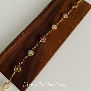 May include: A delicate gold-toned bracelet featuring a series of colorful gemstones. The bracelet is displayed on a wooden surface. The gemstones are various colors, including pink, green, and red. The bracelet is a piece of jewelry.