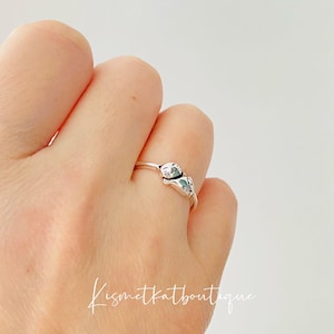 Op de afbeelding: Een zilveren ring met twee kleine kattenfiguren die elkaar aankijken. De ring zit aan een vinger.