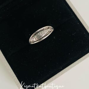 May include: A silver ring with a detailed fish design. The ring is displayed in a black velvet box with a white border and the text "Kismetkatboutique".