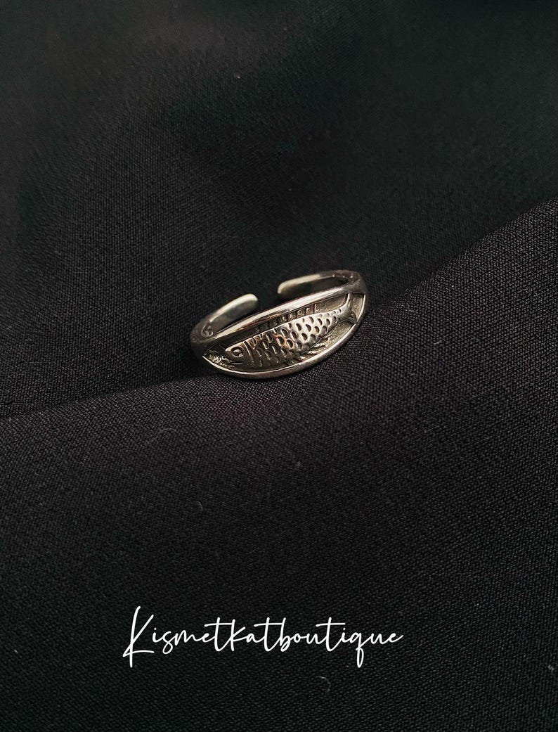 May include: A silver adjustable ring with a fish design. The fish is detailed with scales and has a slightly curved shape.