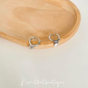 May include: A pair of silver hoop earrings with a small heart charm hanging from each hoop. The earrings are displayed on a wooden surface.