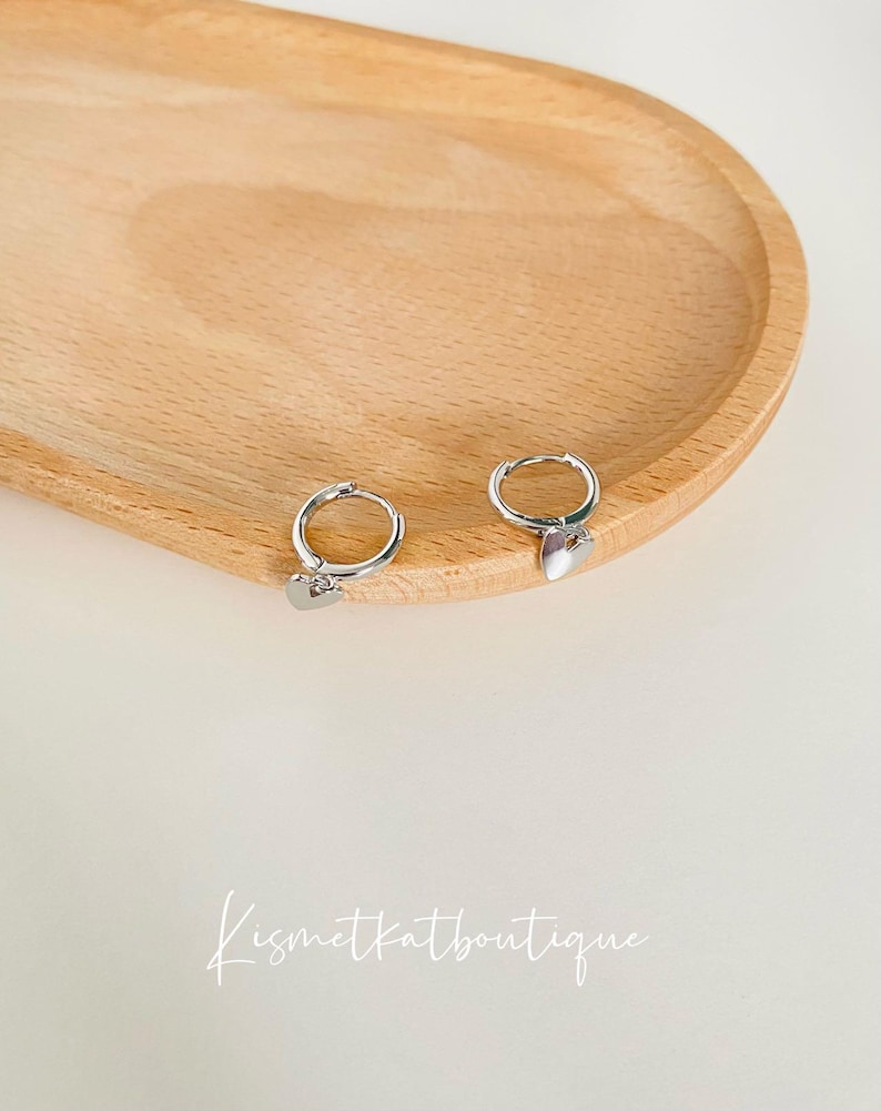 May include: A pair of silver hoop earrings with a small heart charm hanging from each hoop. The earrings are displayed on a wooden surface.