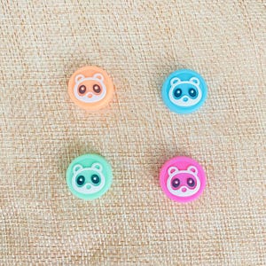 May include: Four round silicone button covers with a cartoon animal face design. The covers are in orange, blue, green, and pink.