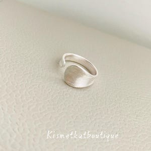 May include: A silver-toned ring with a wide, curved, and textured front panel that tapers into a smooth, open-ended band. The surface of the ring features a brushed metallic finish. The text Kismetkatboutique is visible in the lower portion.