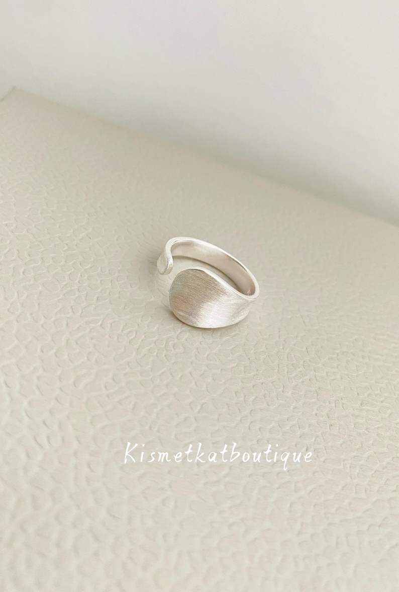 May include: A silver-toned ring with a wide, curved, and textured front panel that tapers into a smooth, open-ended band. The surface of the ring features a brushed metallic finish. The text Kismetkatboutique is visible in the lower portion.