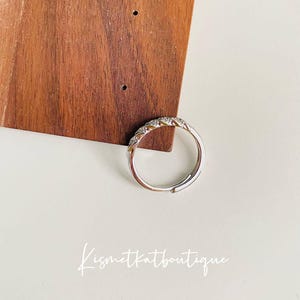 Op de afbeelding: Een zilverkleurige ring met een rij kleine, heldere stenen langs de bovenkant. De ring wordt getoond op een witte ondergrond met een houten achtergrond. De tekst "Kismetkatboutique" is in sierlijke letters geschreven.