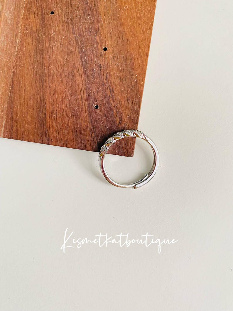Op de afbeelding: Een zilverkleurige ring met een rij kleine, heldere stenen langs de bovenkant. De ring wordt getoond op een witte ondergrond met een houten achtergrond. De tekst "Kismetkatboutique" is in sierlijke letters geschreven.