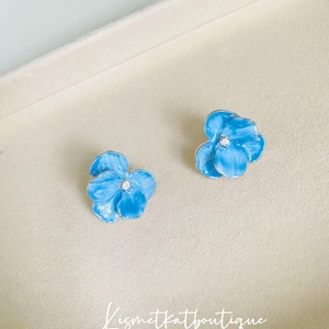Puede incluir: Un par de pendientes de botón de flor azul con un pequeño strass en el centro de cada flor.
