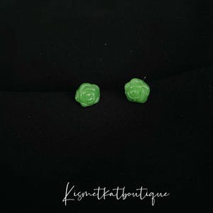 Puede incluir: Un par de pendientes de color verde en forma de rosa. Los pendientes están hechos de un material liso y brillante.
