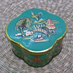 May include: A vintage, four-lobed, teal tin box with a gold trim. The lid features an Asian-inspired landscape with a bridge, pagoda, and boat. The box is likely for storage or decoration. The box is sitting on a woven mat.