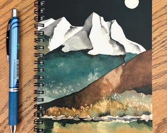 May include: A spiral-bound notebook featuring a watercolor landscape of snow-capped mountains under a full moon against a black sky, with a blue retractable pen positioned to the left. The artist's signature, Kathy Cooney, is in the bottom right.