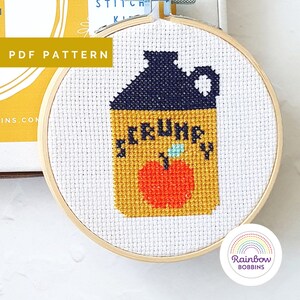 Könnte beinhalten: Ein Kreuzstichmuster eines gelben Glases mit schwarzem Deckel und einem roten Apfel. Das Glas trägt die Aufschrift "CRUMPY" auf der Vorderseite.