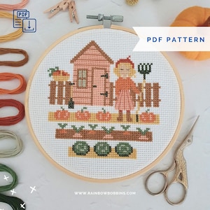 May include: A cross-stitch project in a wooden hoop featuring a garden scene with a pink shed, a person holding a garden tool, pumpkins, carrots, and leafy greens. The design includes the text PDF PATTERN. Embroidery floss and scissors are nearby.