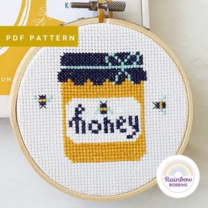May include: A white embroidery hoop with a cross-stitch design of a honey jar with a bee and the word "honey" on the label. Two bees are also stitched on the background.