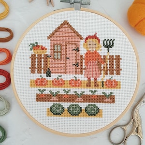 May include: A cross-stitch embroidery project in a wooden hoop featuring a garden scene with a pink shed, a person in a red jumper, pumpkins, carrots, and leafy greens. The design is stitched on white Aida cloth with colourful embroidery floss.