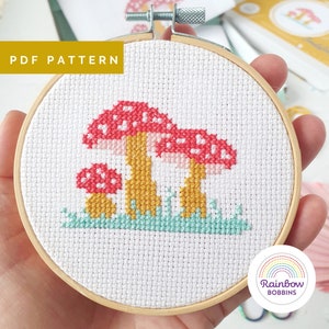 May include: A cross-stitch embroidery of mushrooms in a wooden hoop. The mushrooms have red caps with white spots and yellow stems, set against a white background. The text "PDF PATTERN" is visible. The design includes green grass and is from Rainbow Bobbins.