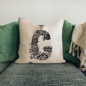 K&ouml;nnte beinhalten: Ein wei&szlig;es Kissen mit einer schwarzen und wei&szlig;en Kritzelei des Buchstabens "G" darauf. Das Kissen liegt auf einem gr&uuml;nen Sofa.