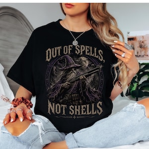 May include: A black short-sleeved t-shirt featuring a graphic print of a wizard in a purple robe holding a shotgun. The text "OUT OF SPELLS, NOT SHELLS" is printed in a distressed, tan-coloured font above and below the central circular illustration.