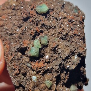 Op de afbeelding: Een close-up van een ruwe, bruine rots met verschillende kleine, groene, kristalvormige formaties. De rots is bedekt met kleine, witte vlekken.