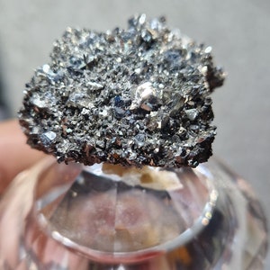 Può includere: Un ammasso di piccoli cristalli metallici grigi con un bagliore argenteo. I cristalli sono disposti in una forma irregolare e ruvida e sono esposti su un piedistallo di vetro trasparente.