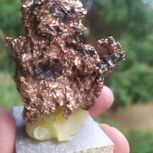 May include: A close-up of a rough, metallic, copper-colored rock on a small wooden base. The rock has a unique, irregular shape and a textured surface.