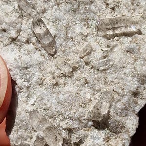 Puede incluir: Primer plano de una roca con varios pequeños cristales de cuarzo transparentes incrustados. Los cristales son puntiagudos y tienen un aspecto vítreo.