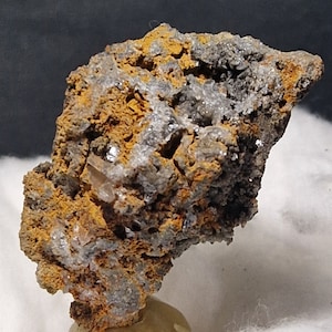 Könnte beinhalten: Ein rauer, brauner und grauer Stein mit einem Cluster aus kleinen, klaren Kristallen. Der Stein liegt auf einer weißen, flauschigen Oberfläche.