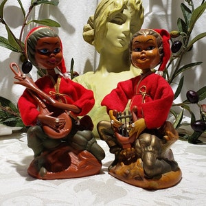 May include: Two ceramic figurines of elves playing musical instruments. The elf on the left is wearing a red and gold outfit and playing a lute. The elf on the right is wearing a red and gold outfit and playing a harp. Both elves have brown skin and black hair.