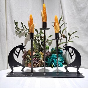 May include: A black metal candle holder featuring three vertical candle holders with corn cobs. The base has two Kokopelli figures on each side. The background includes a floral arrangement with green and purple flowers.