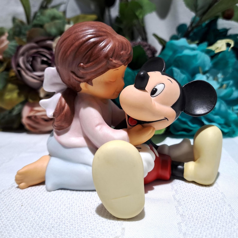 Pode incluir: Uma estatueta de porcelana retratando uma menina com uma camisa rosa e saia azul abra&ccedil;ando uma figura do Mickey Mouse. A menina tem cabelo castanho e um la&ccedil;o branco. Mickey Mouse usa cal&ccedil;&otilde;es vermelhos e sapatos amarelos. Em um fundo floral.