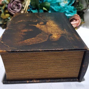 May include: A decorative wooden box designed to resemble a vintage book. The box features a dark brown and gold painted design on the top, with a detailed image. The edges and sides are painted to look like aged book pages.