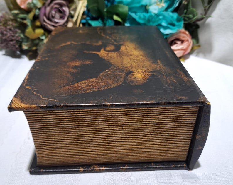 May include: A decorative wooden box designed to resemble a vintage book. The box features a dark brown and gold painted design on the top, with a detailed image. The edges and sides are painted to look like aged book pages.