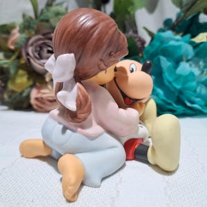 Pode incluir: Uma estatueta de cer&acirc;mica de uma menina com cabelo castanho e um la&ccedil;o branco, abra&ccedil;ando um c&atilde;o de desenho animado. A menina usa uma camisa rosa e cal&ccedil;as azul claro. O c&atilde;o tem um rosto castanho, nariz preto e l&iacute;ngua vermelha. A estatueta est&aacute; em uma superf&iacute;cie branca.