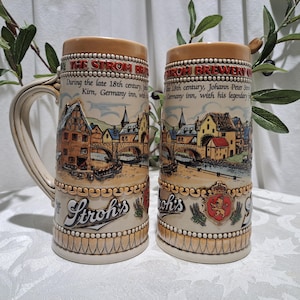 May include: Two ceramic beer steins with a white background and brown, red, and yellow accents. The steins feature a scene of a German village with a bridge and a river. The text "The Stroh Brewery" and "Stroh's" is visible on the steins.