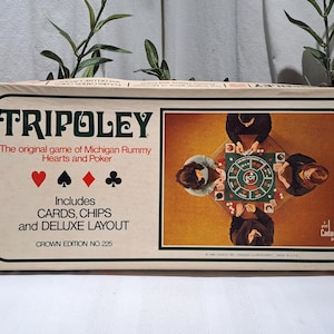 May include: A rectangular Tripoley board game box with a cream-colored background. The front features the title, the text The original game of Michigan Rummy Hearts and Poker, card suit symbols, and an illustration of people playing the game.