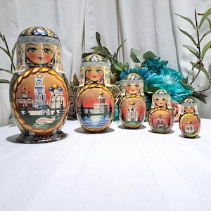 May include: A set of five Russian nesting dolls, each painted with a different scene of buildings and landscapes. The dolls are in shades of brown, orange, and green, with detailed facial features and decorative headwear. The largest doll has text on the bottom.