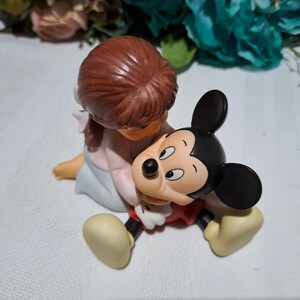 Pode incluir: Uma estatueta de uma crian&ccedil;a abra&ccedil;ando uma figura do Mickey Mouse. A crian&ccedil;a tem cabelo castanho e est&aacute; vestida com uma roupa rosa claro e azul. Mickey Mouse est&aacute; com sua roupa cl&aacute;ssica preta, vermelha e amarela.