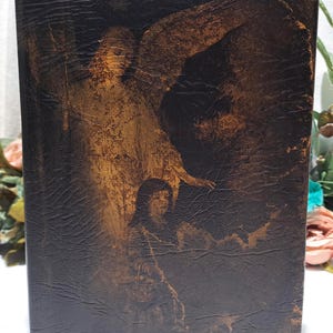 May include: A vintage book with a dark brown, textured cover. The cover features a sepia-toned illustration of an angel and a person. The artwork is detailed, with visible brushstrokes and shading, creating a classic, antique aesthetic.