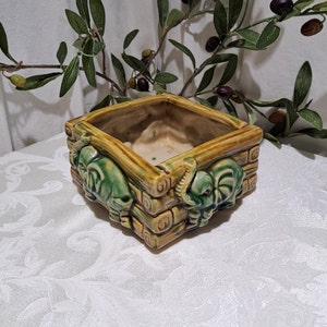 May include: A square ceramic planter with a green and brown glaze. The planter is decorated with three elephant figures on the sides. The planter is empty.