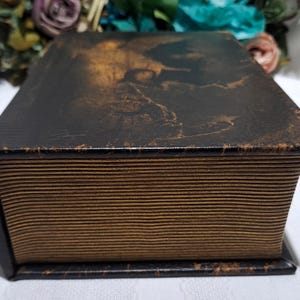 May include: A vintage-style, rectangular box with a dark brown, textured exterior. The box has a lid with a faded, sepia-toned image. The sides of the box have a ribbed, paper-like appearance, and the edges are dark brown.