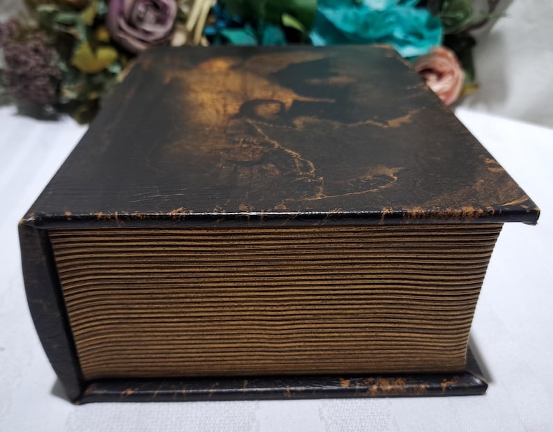 May include: A vintage-style, rectangular box with a dark brown, textured exterior. The box has a lid with a faded, sepia-toned image. The sides of the box have a ribbed, paper-like appearance, and the edges are dark brown.